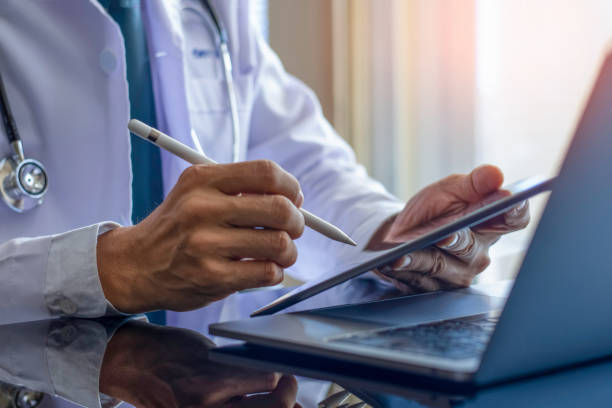 Male doctor in white coat with  medical stethoscope, hand holding and using modern digital tablet, work on laptop computer at office. Telehealth, Online medical networking concept.