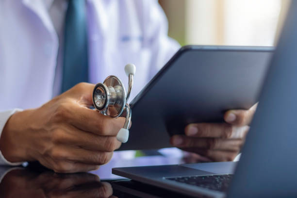 Male doctor in white lab coat, hand holding medical stethoscope and using modern digital tablet pc, 
work on laptop computer at workplace. Online medical, e health or medical network concept.